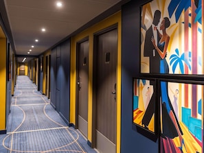 Un pasillo de hotel con puertas a la derecha, una alfombra estampada y un mural colorido en la pared.
