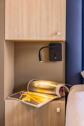 A wooden bedside table with a magazine, a wall-mounted lamp, and a power outlet. - Kyriad Saint Quentin (Saint-Quentin)