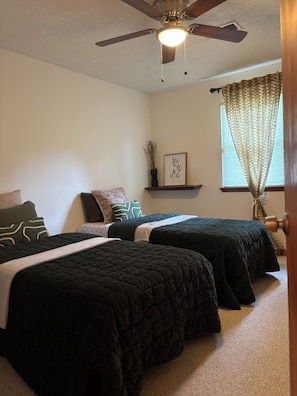 A bedroom with two beds, a ceiling fan, a window with curtains, and a small shelf with a plant and picture.