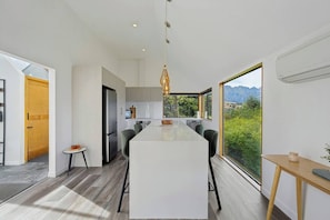 A modern kitchen with a central island, large windows offering a mountain view, and a dining area with a long table. - Earnslaw Lakeview Retreat, close to town + parking (Queenstown)