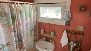 A bathroom with a shower curtain featuring palm trees and cars, a sink, a window with a mirror, and a sign that says "Relax! You're on beach time." - Charming 1-bedroom cottage in York perfect for a relaxing getaway. (York)