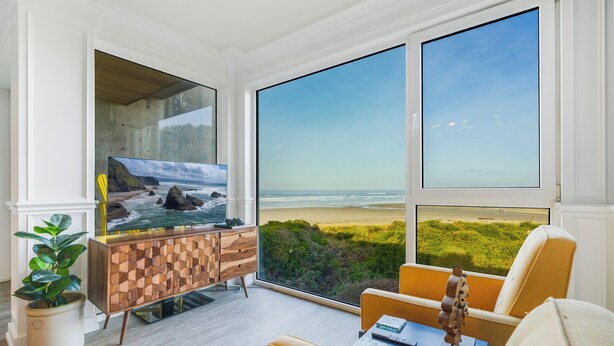 A room with a large window overlooking a beach, a wooden cabinet, a plant, and a table with books. - Rockaway Beach Oceanfront Views! Private Balcony, Beachfront Paradise! (Rockaway Beach)