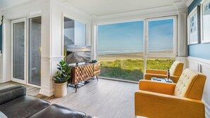 A spacious room with a large window offering a view of the beach, a wooden cabinet, a leather sofa, and a yellow armchair. - Rockaway Beach Oceanfront Views! Private Balcony, Beachfront Paradise! (Rockaway Beach)
