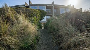 A modern house with a curved roof, surrounded by dense vegetation and a sandy path leading to the entrance. - Rockaway Beach Oceanfront Views! Private Balcony, Beachfront Paradise! (Rockaway Beach)