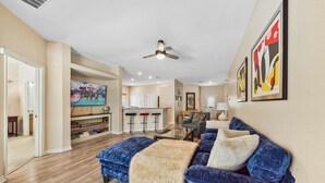 A spacious living room with a blue sofa, a glass coffee table, and a ceiling fan. There is a kitchen area with a bar and stools in the background. - Epic Princess and Villains Hideaway-  Private Pool  Themed Rooms  Near Disney (Kissimmee)