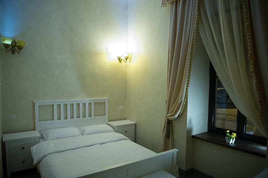 Un dormitorio con cama, mesitas de noche, una lámpara de pared, una ventana con cortinas y vistas al exterior.