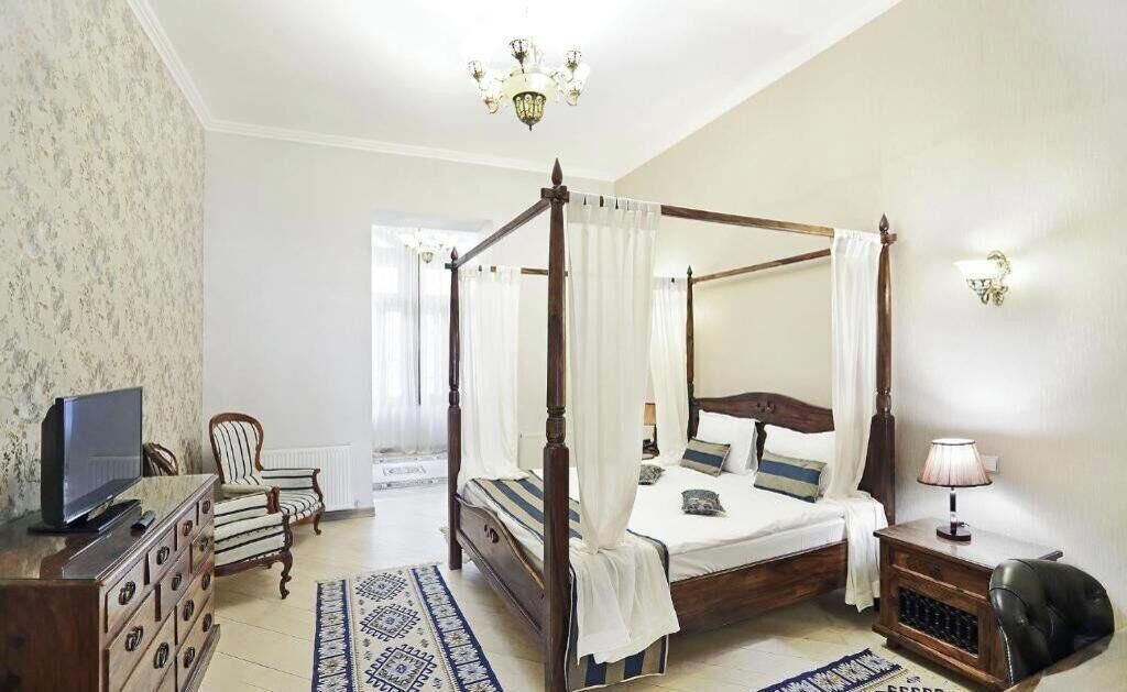 A four-poster bed with white curtains, a television on a wooden dresser, a patterned rug, and a lamp on a side table.