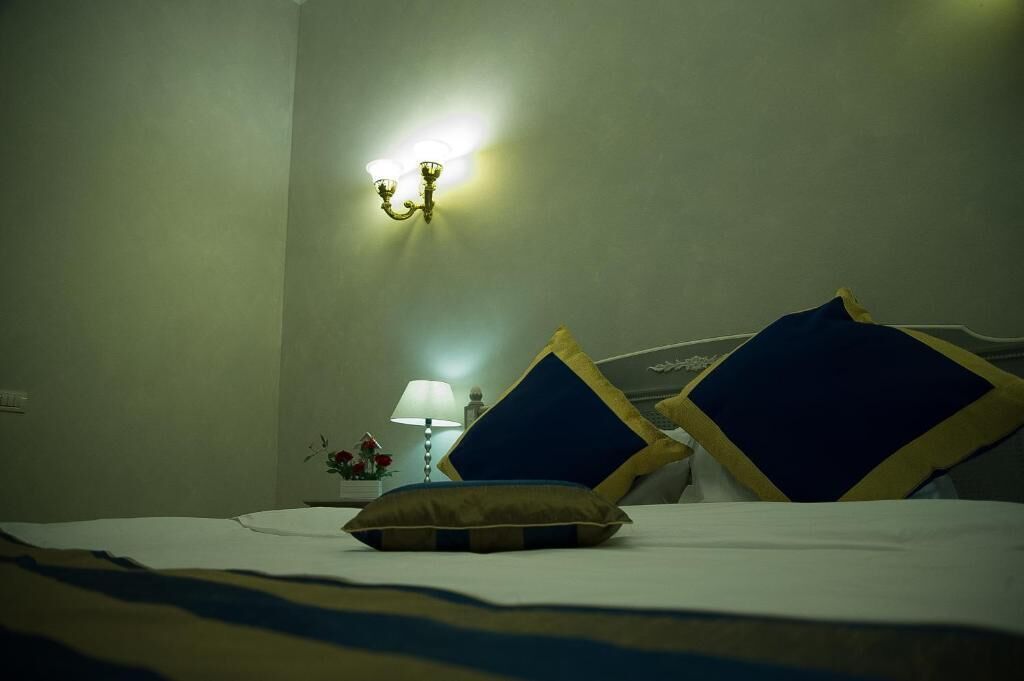 A hotel room with a bed, pillows, a nightstand with a lamp, and a wall-mounted light fixture.