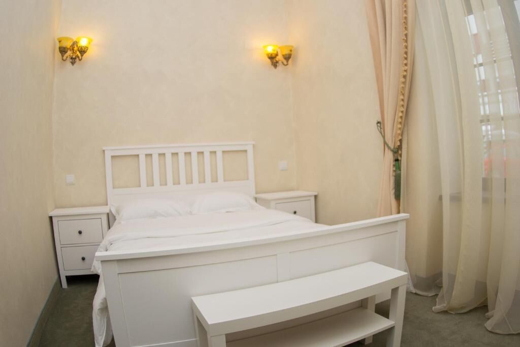 A white bed with a headboard, two bedside tables, a bench, and wall-mounted lights.
