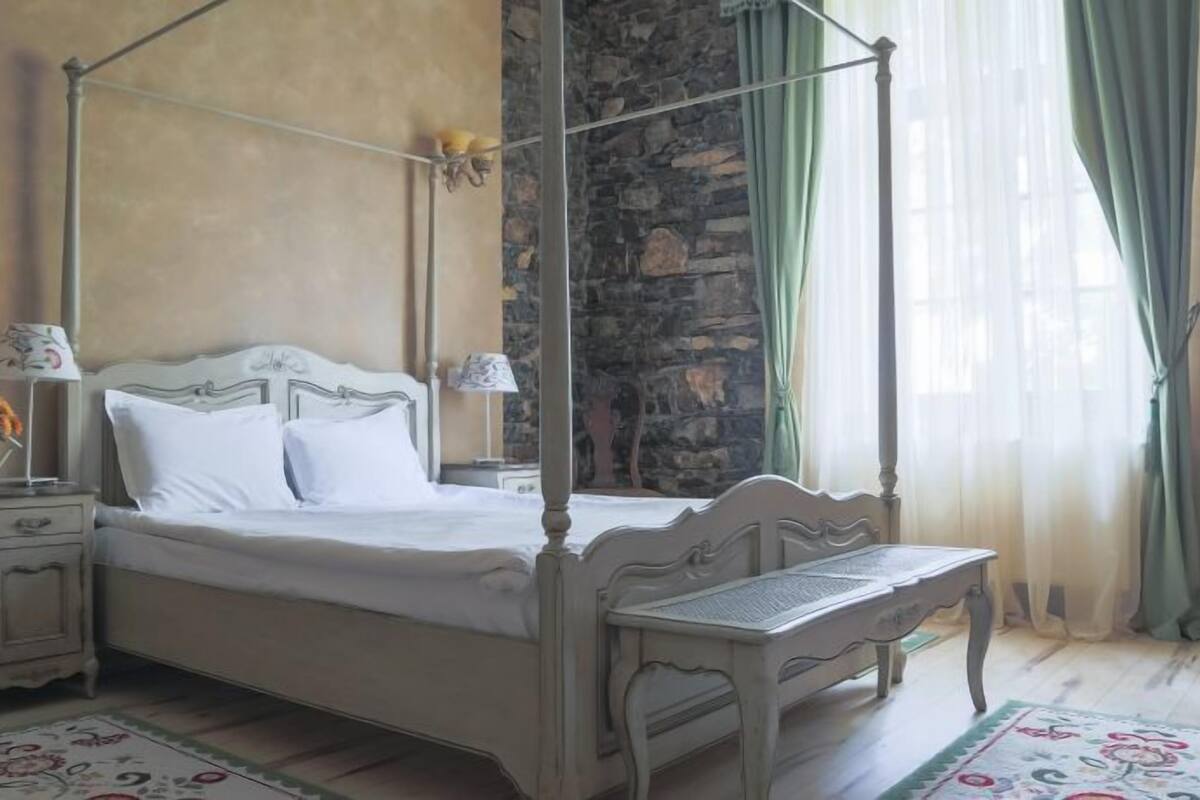 A four-poster bed with a stone wall backdrop, a bedside table with a lamp, and a floral-patterned rug.