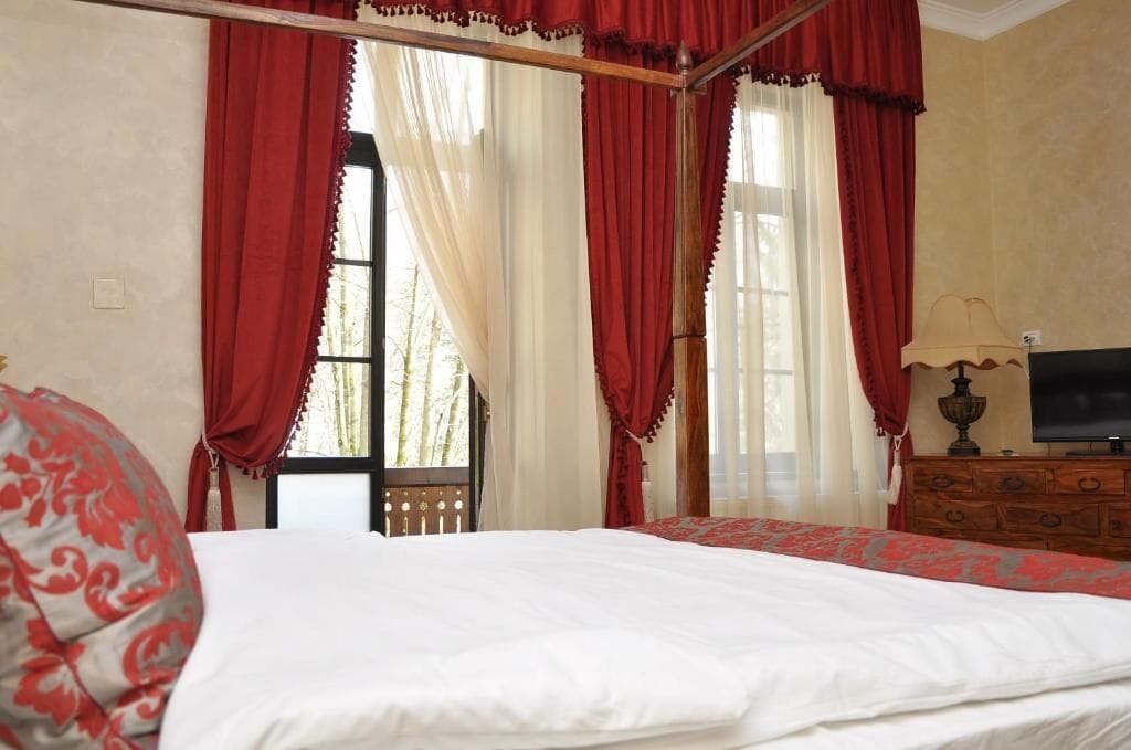 A bedroom with a four-poster bed, a television on a wooden dresser, and a window with red curtains.