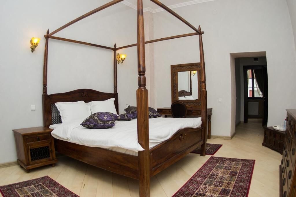 Una cama con dosel de madera, ropa de cama blanca y almohadas moradas. Hay dos mesitas de noche con lámparas, un espejo en la pared y una alfombra estampada en el suelo.