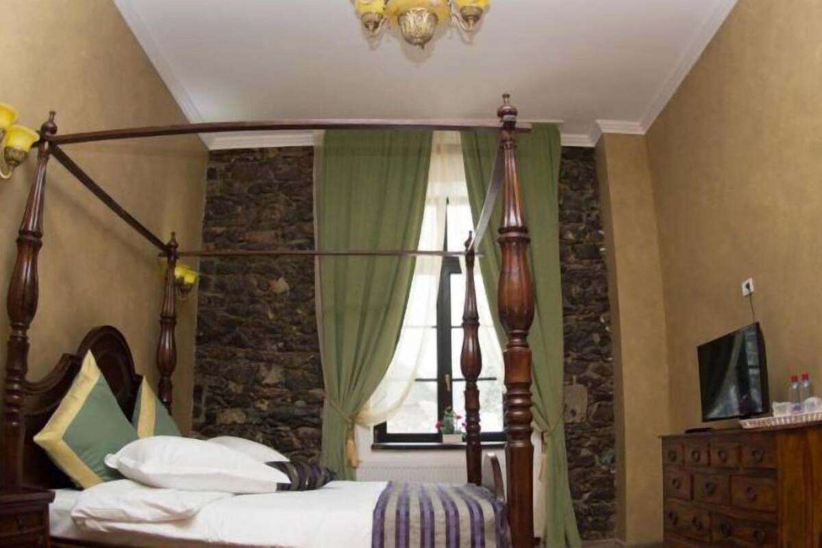 A bedroom with a four-poster bed, a stone wall, a window with curtains, a dresser, and a television.