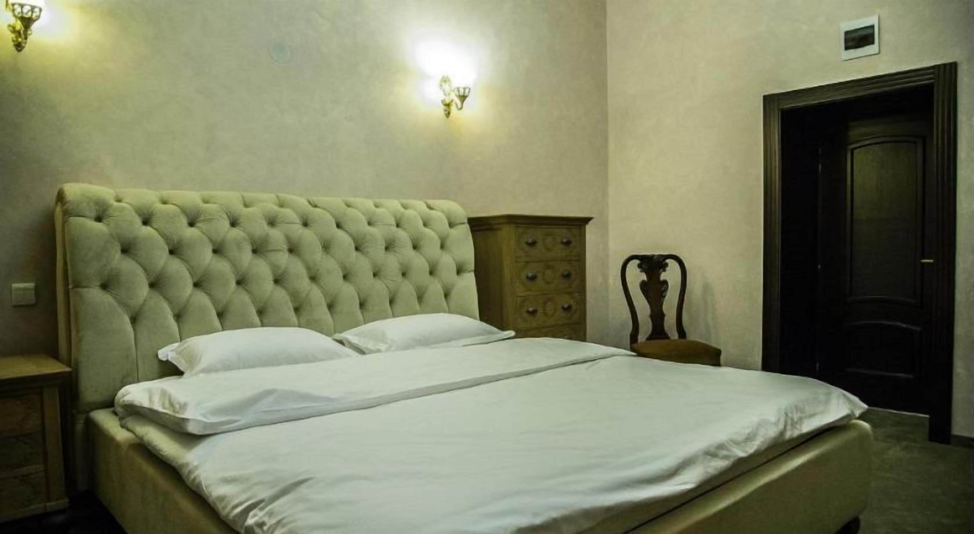 Un lit bien fait, doté d’une tête de lit capitonnée, une table de chevet en bois avec une lampe, et une porte sombre dans une pièce aux murs de couleur claire.