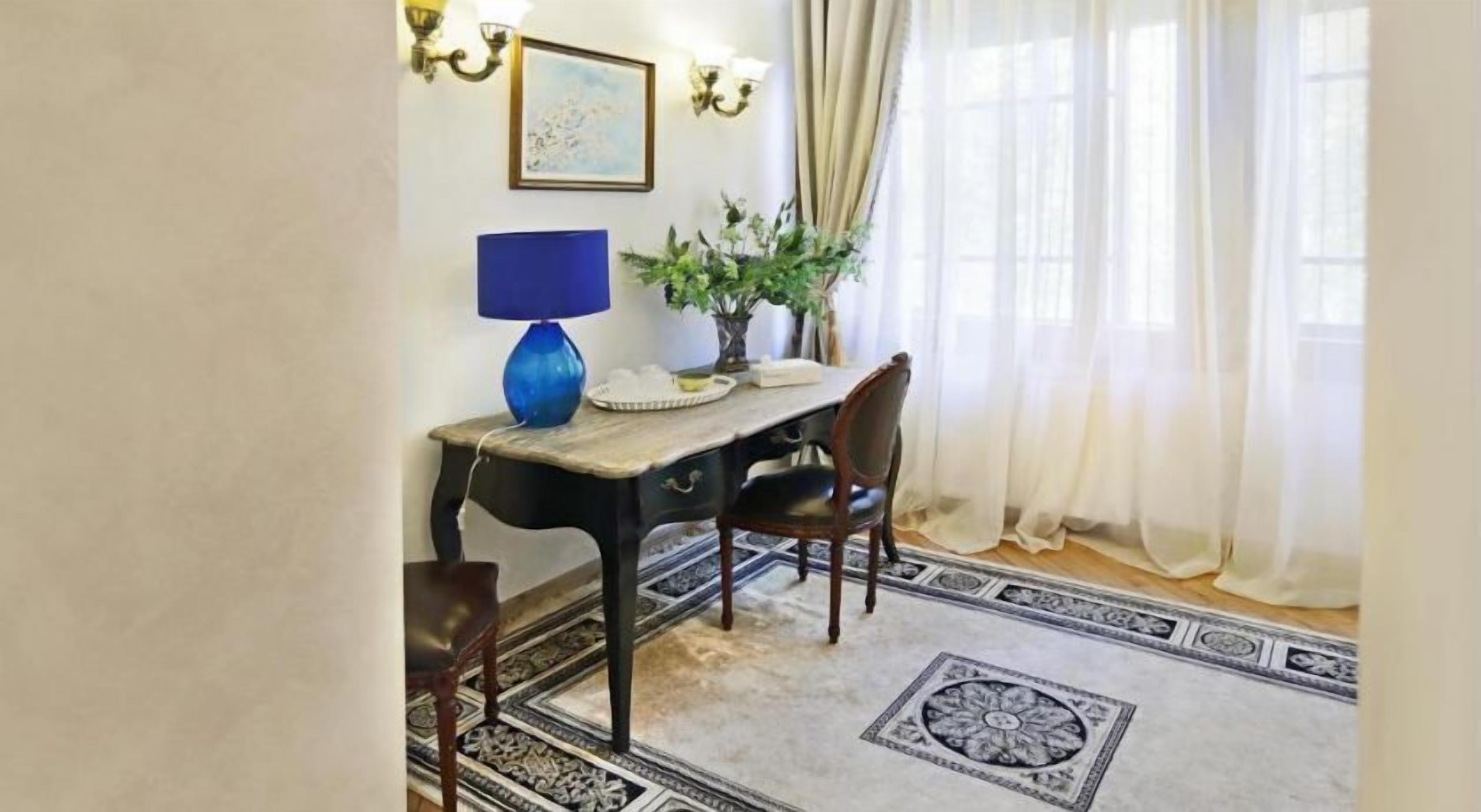 A dining area with a table, chairs, a lamp, a plant, and a patterned rug.