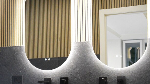 Ein Badezimmer mit zwei großen, runden Steinswaschbecken, einem Holzwaschtisch und einem Spiegel mit einem ungewöhnlichen, geschwungenen Design.