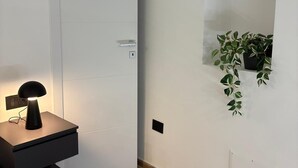 Ein modernes Schlafzimmer mit weißer Tür, einem Nachttisch mit Lampe, einer kleinen Pflanze und einem Bett mit strukturierter Bettdecke.