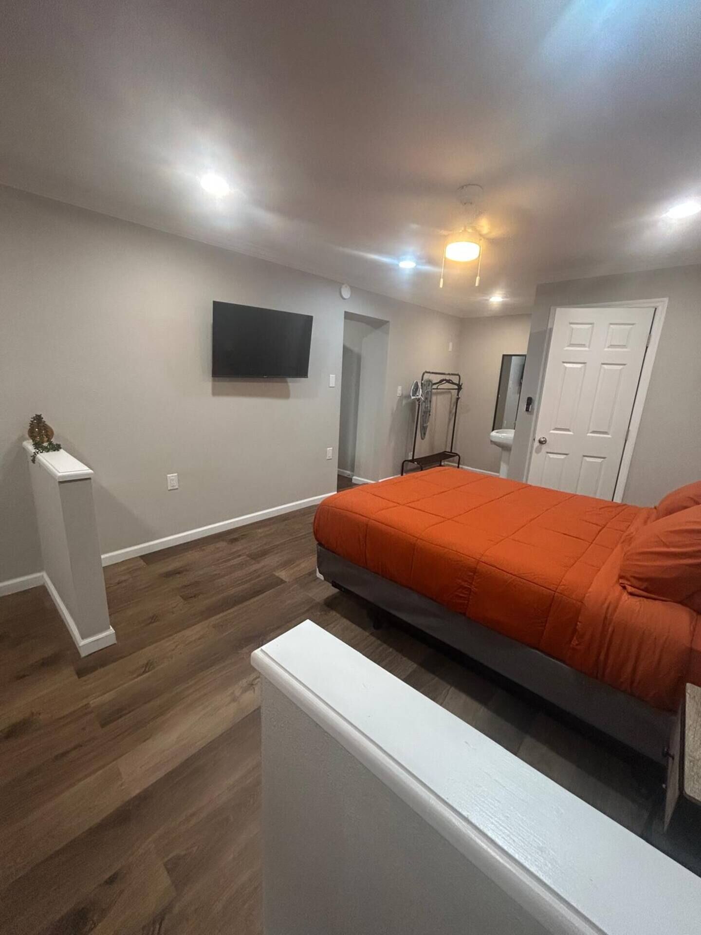 A bedroom with a bed, a TV mounted on the wall, a door, and a small bathroom area with a mirror and sink.