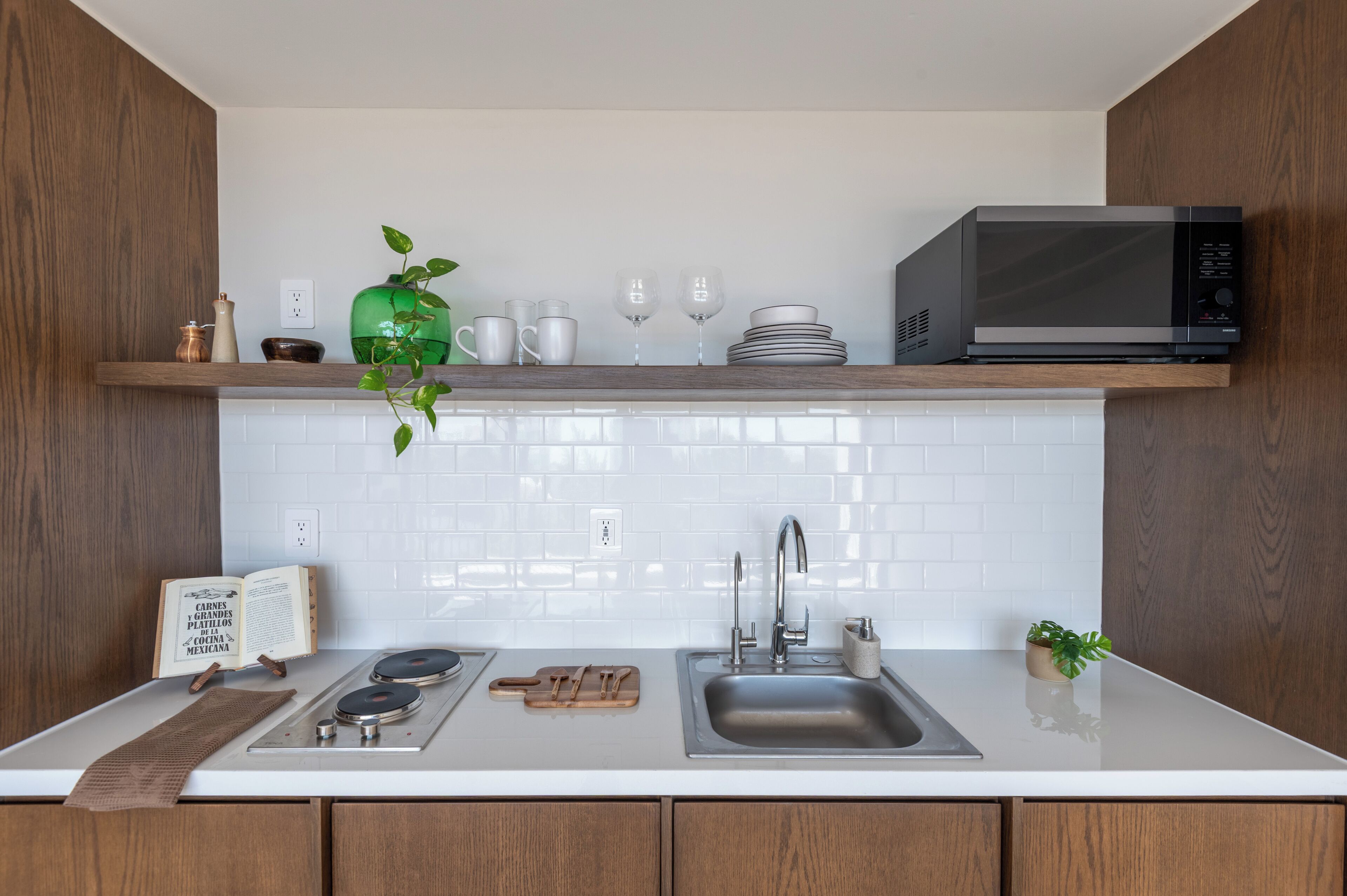 Uma cozinha moderna com bancada branca, pia de aço inoxidável e micro-ondas. Há uma prateleira de madeira acima do balcão com um vaso verde, xícaras brancas e uma pilha de pratos.