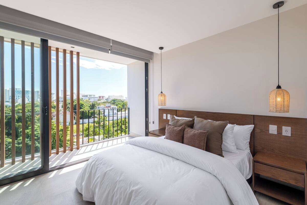 Une chambre moderne avec une grande fenêtre, un lit recouvert d’une literie blanche et deux lampes suspendues.