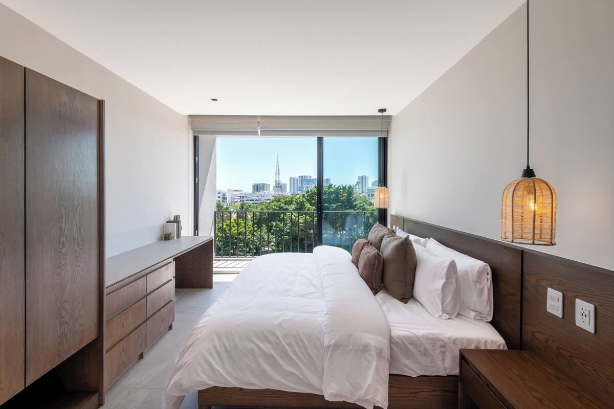 Une chambre moderne avec un grand lit, une commode en bois, une table de chevet et une vue sur les toits de la ville, visible à travers de grandes fenêtres.
