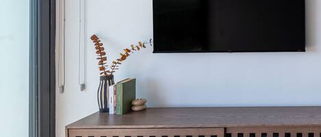 A modern living room with a large flat-screen TV mounted on a white wall, a wooden bench with storage, and a large window providing natural light.