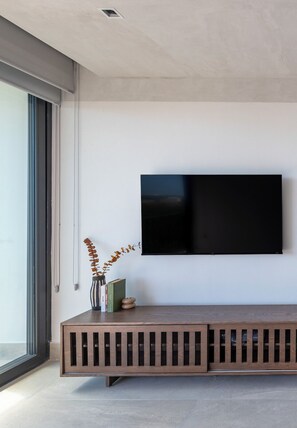 Ein modernes Wohnzimmer mit einem großen Flachbildfernseher an einer weißen Wand, einer Holzlounge mit Stauraum und einem großen Fenster, das natürliches Licht hereinlässt.