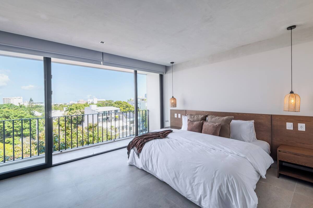 Une chambre moderne dotée d’une grande fenêtre, d’un lit avec une literie blanche, d’une tête de lit en bois et d’un luminaire suspendu.