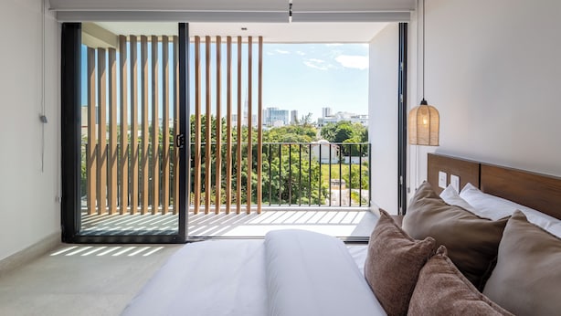 A bedroom with a large bed, a view of a cityscape through a sliding glass door, and a balcony with a wooden railing.