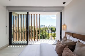 A bedroom with a large bed, a view of a cityscape through a sliding glass door, and a balcony with a wooden railing. - Mojarra 27 New Apts Heart of Cancun (CancΓΊn)