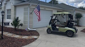 A residential house with a garage, an American flag, a golf cart, and a palm tree in the driveway. - The Villages 2 bedroom / 2 bath home minutes from Lake Sumter Landing (The Villages)