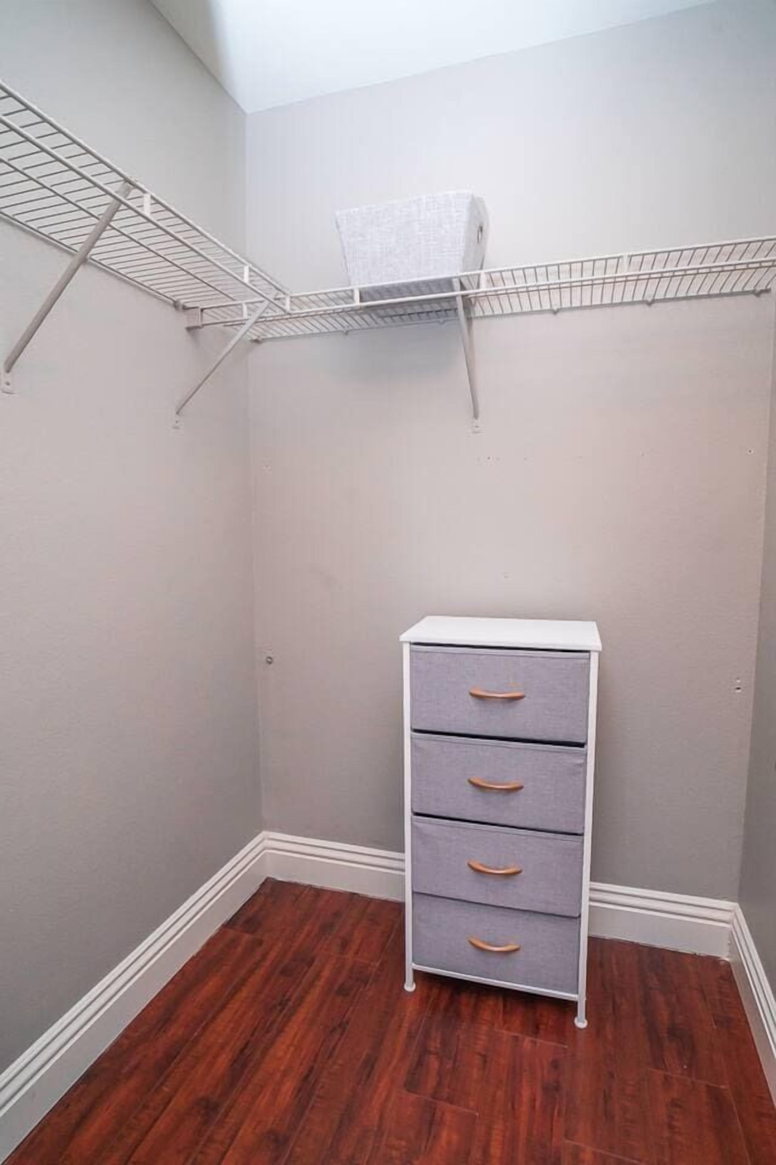 Ein kleines Zimmer mit Holzboden, einer weißen Kommode mit Schubladen und einem Drahtregal darüber.