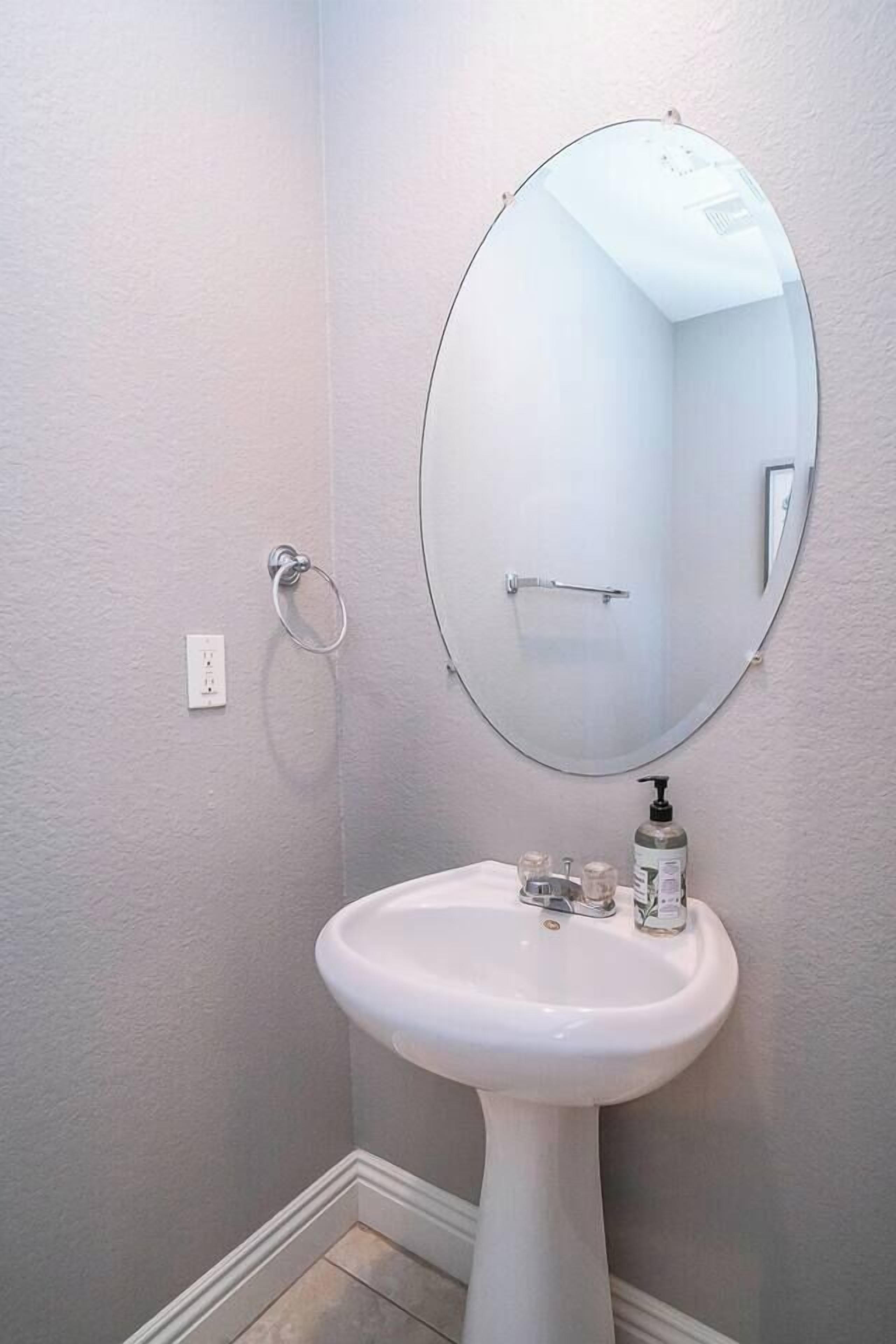 Une salle de bain avec un lavabo sur colonne blanc, un miroir rond et un distributeur de savon liquide.
