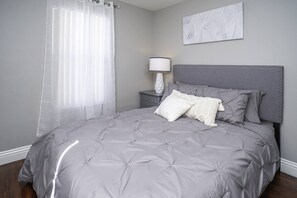 Een slaapkamer met een grijs gestoffeerd bed, een nachtkastje met een lamp en een kunstwerk aan de muur.