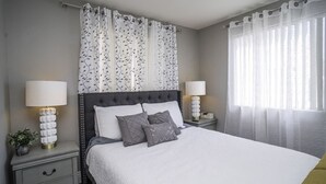 A neatly arranged bedroom with a tufted headboard, a large bed with white bedding, two bedside tables with lamps, and sheer curtains on the windows.