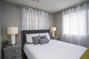 A neatly arranged bedroom with a tufted headboard, a large bed with white bedding, two bedside tables with lamps, and sheer curtains on the windows. - 3BR Home Near Las Vegas Strip | Pet Friendly (Las Vegas)