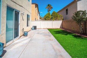 Un jardin arrière d’une maison, avec une terrasse, une pelouse, une clôture blanche et un palmier.