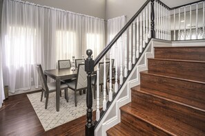 A dining area with a table and chairs, a staircase with wooden steps and white balusters, and a patterned rug on the floor.