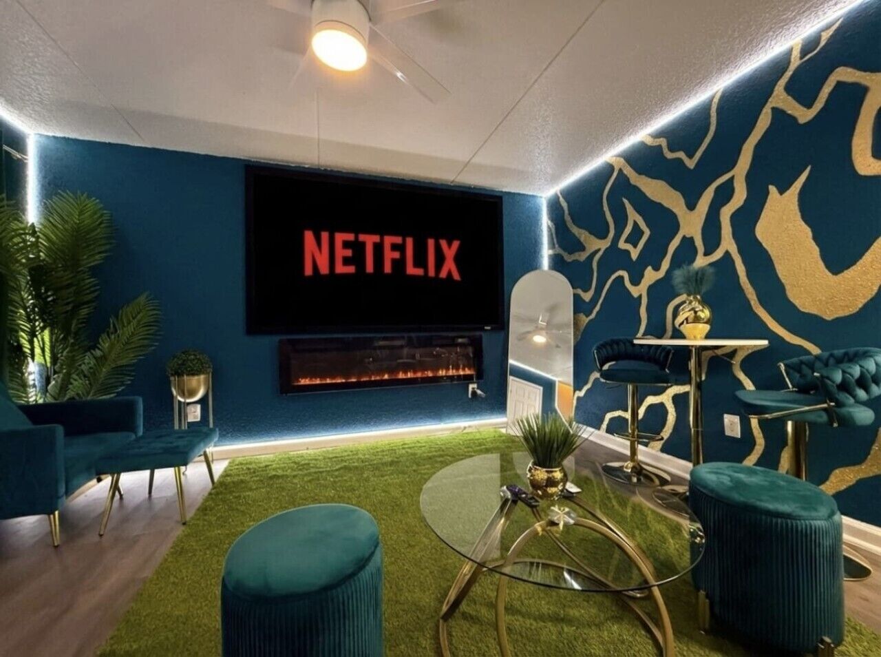 A modern living room with a large flat-screen TV displaying 'NETFLIX,' a fireplace, teal furniture, and a patterned wall.