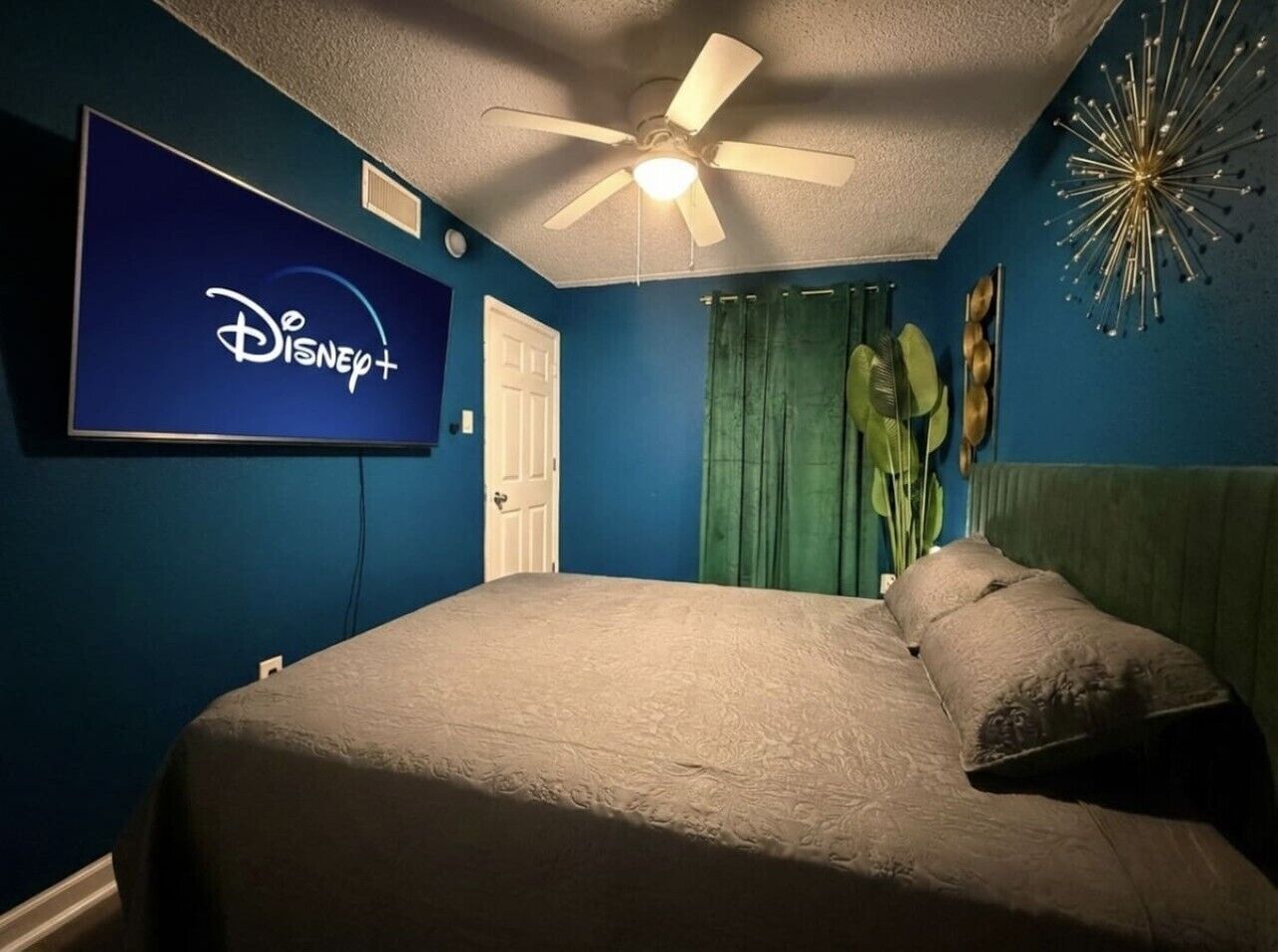 Un dormitorio con cama, ventilador de techo, televisor de pared mostrando el logo de Disney+ y cortina verde.