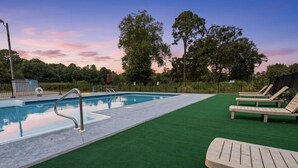 Piscina com escada de metal, rodeada por grama sintética verde e espreguiçadeiras de madeira.
