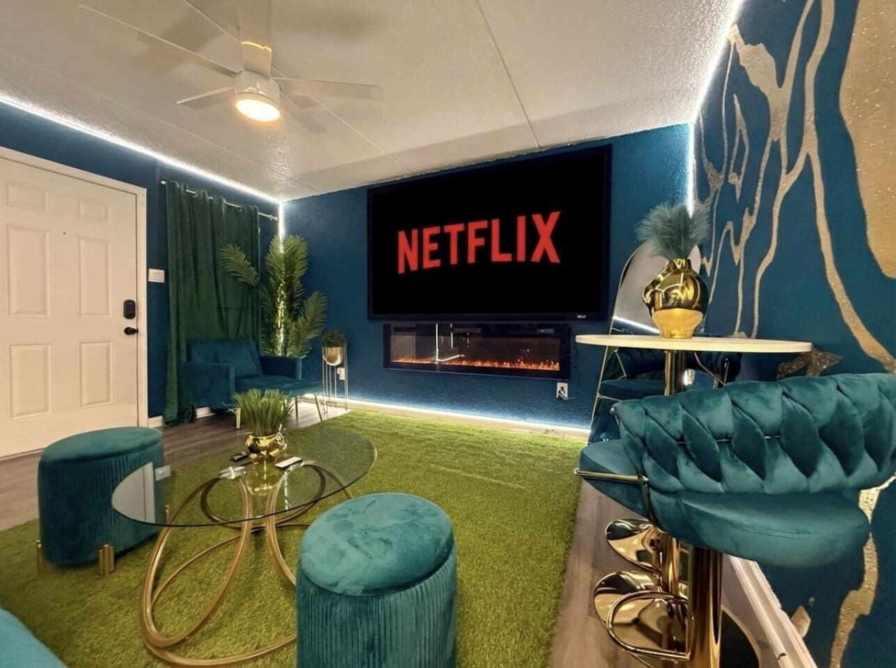 Ein modernes Wohnzimmer mit einem Flachbildfernseher, der 'NETFLIX' anzeigt, einem Kamin, türkisfarbenen Möbeln und einem Deckenventilator.
