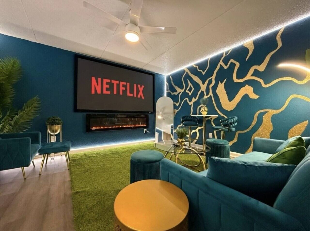 A modern living room with a large flat-screen TV displaying 'NETFLIX,' a fireplace, teal sofas, and a decorative wall with a gold pattern.
