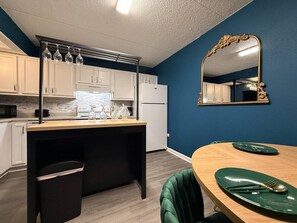 Et kompakt køkken med hvide skabe, en træbordplade og et spiseområde med et grønt bord og stole.