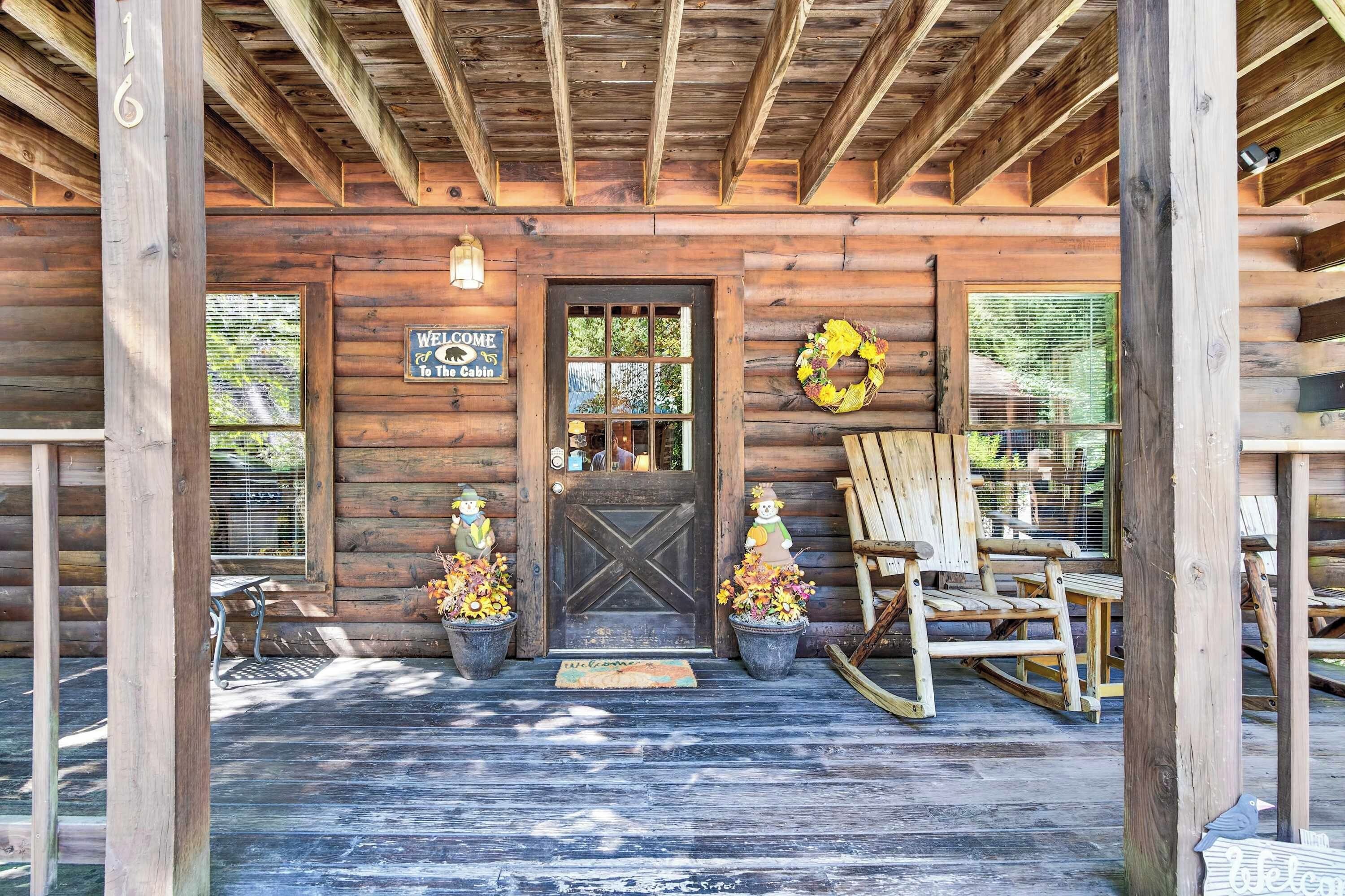 A wooden cabin with a dark wooden door, a welcome sign, two potted plants with autumn flowers, a rocking chair, and a birdhouse.