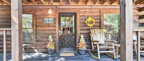 A wooden cabin with a dark wooden door, a welcome sign, two potted plants with autumn flowers, a rocking chair, and a birdhouse.