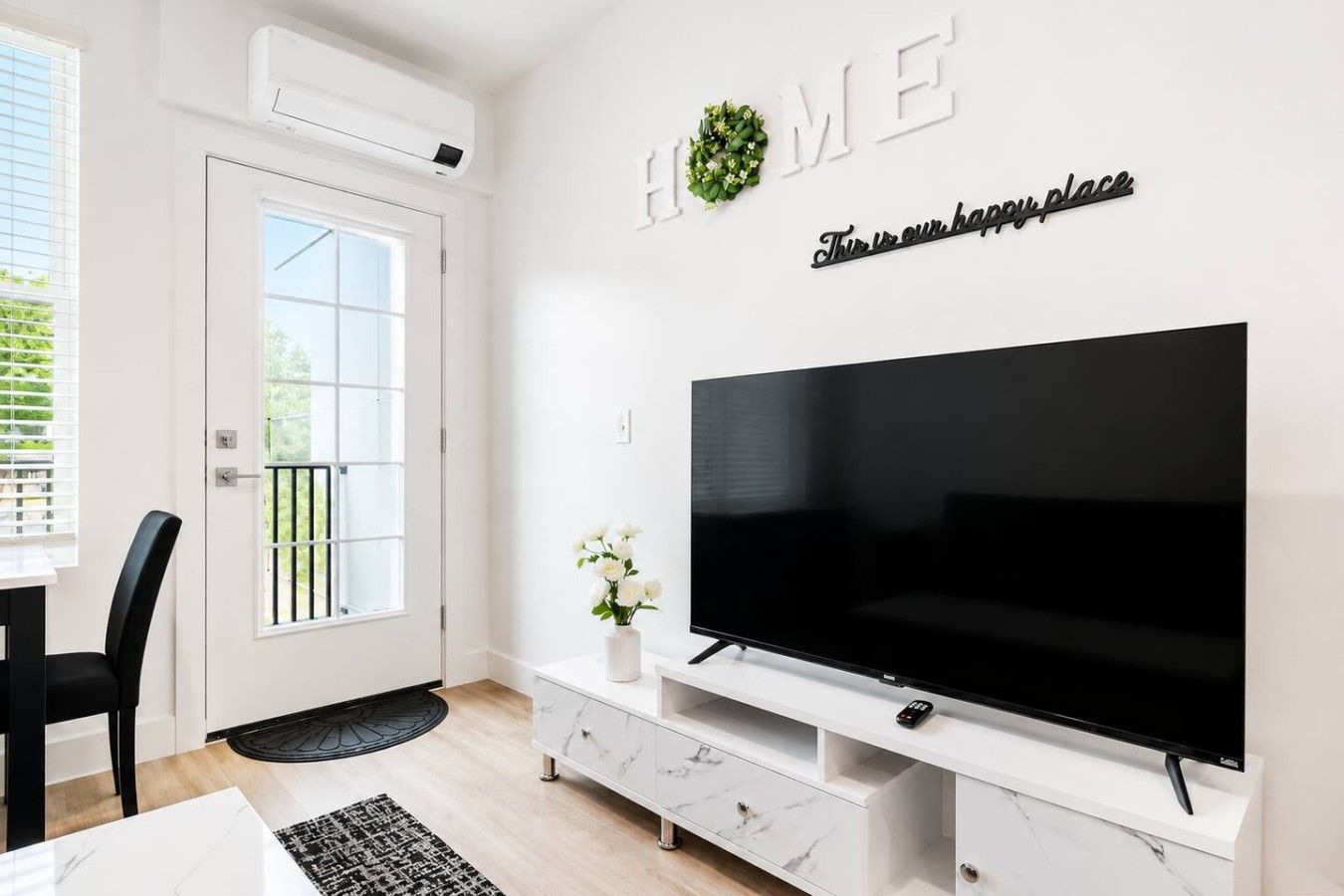 Een moderne woonkamer met een grote flatscreen-tv, een witte tv-kast, een deur met raam en een bordje met de tekst 'HOME' erboven.