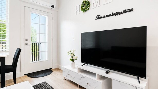 A modern living room with a large flat-screen TV, a white entertainment unit, a door with a window, and a 'HOME' sign above the TV. - Charming 1 BR • Balcony • Free Parking • Downtown (Hot Springs)