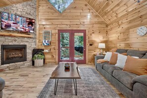 A cozy cabin interior with a stone fireplace, a television, a wooden ceiling, and a large window overlooking trees. - Free Tickets,Sauna,Cold Plunge,Gatlinburg, 1 level (Gatlinburg)