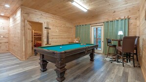 Een houten interieur met een pooltafel, stoelen, een tafel met een lamp en een deur naar een opslagruimte.